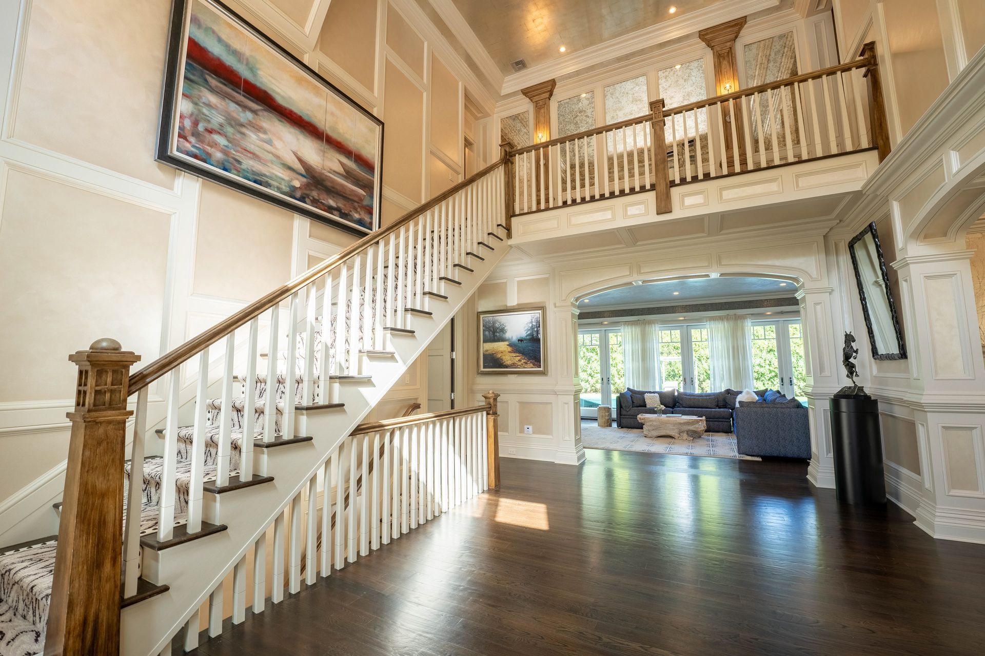 A grand foyer featuring a sweeping staircase with white spindles, dark wood floors, and a view into a living room.
