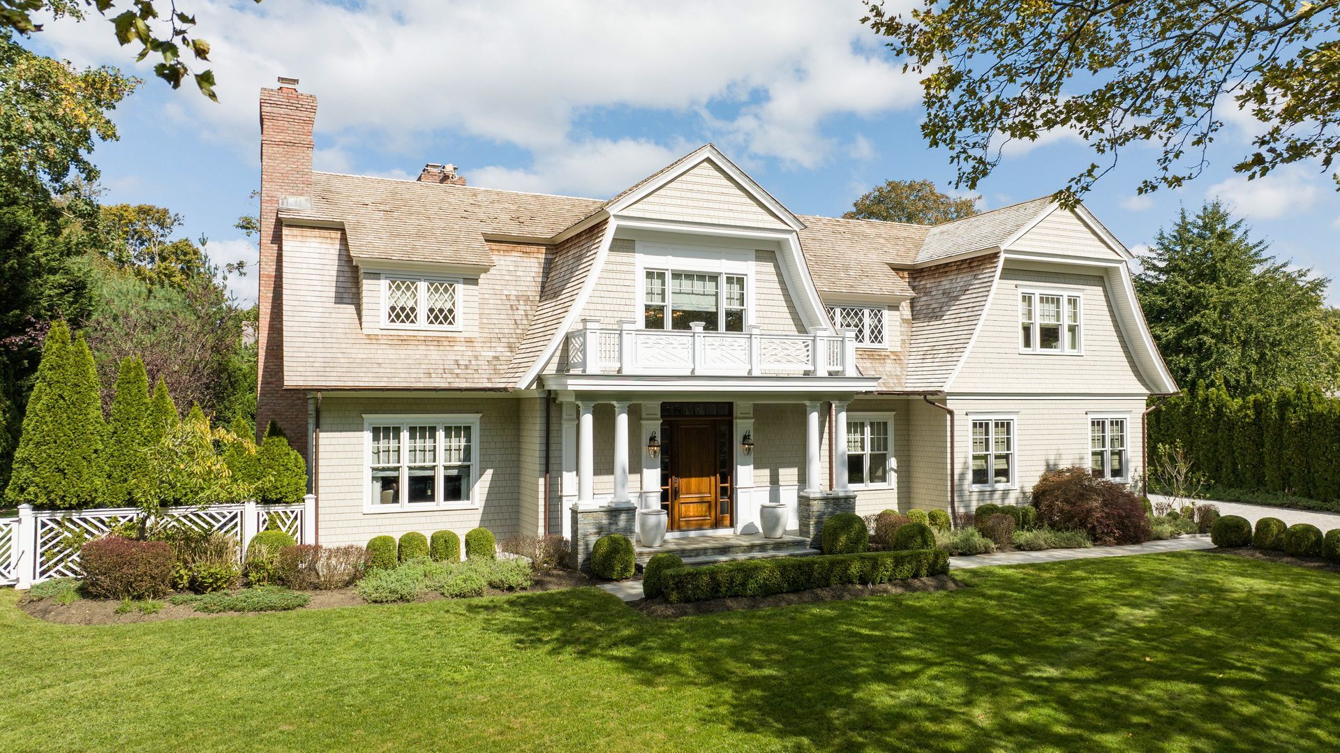 A light-colored, two-story Dutch Colonial house with cedar shingles, a balcony, and a green lawn under a sunny, blue sky.