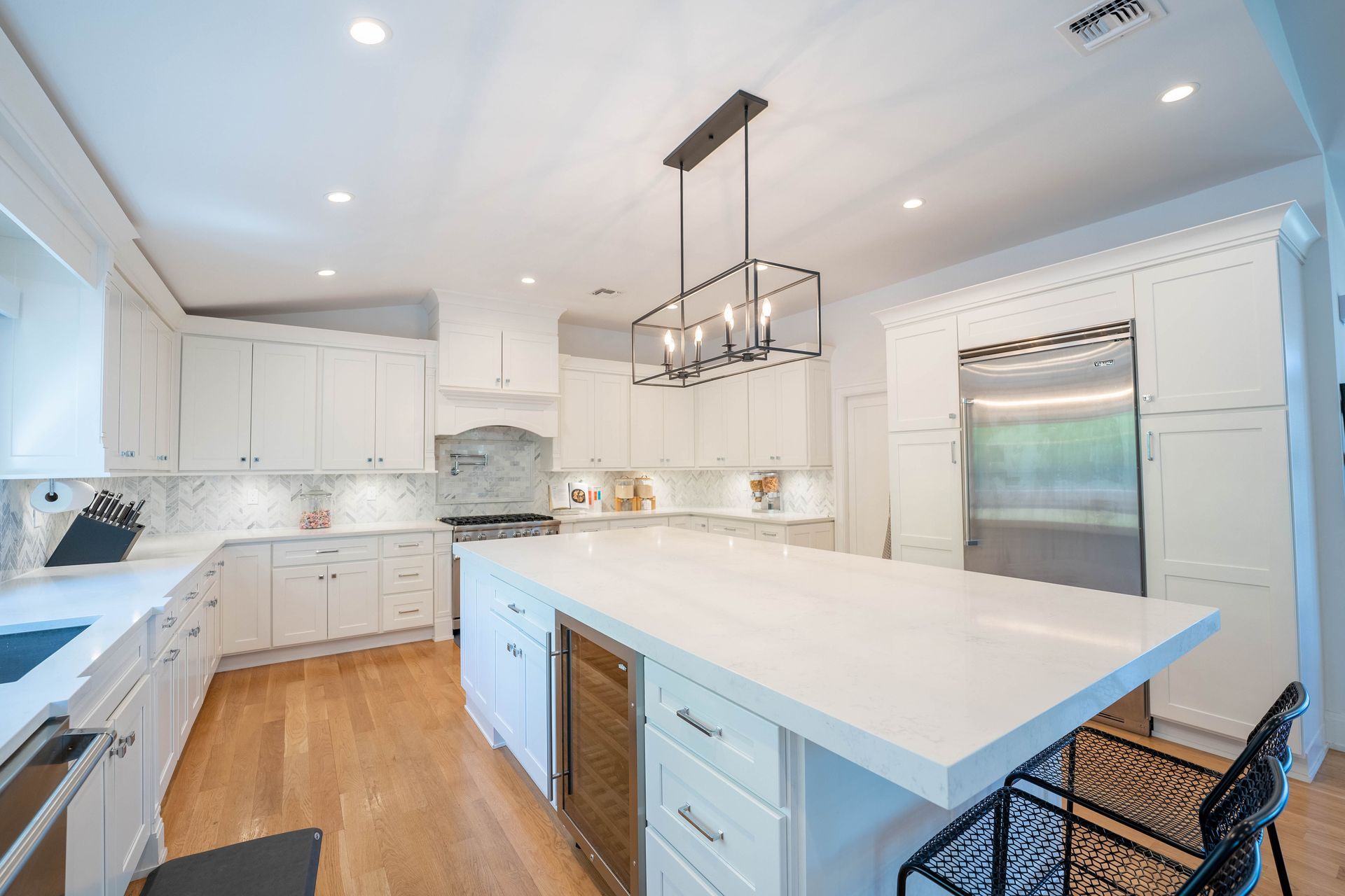 Modern white kitchen with a large island, wood flooring, stainless steel refrigerator, and hanging light fixture.