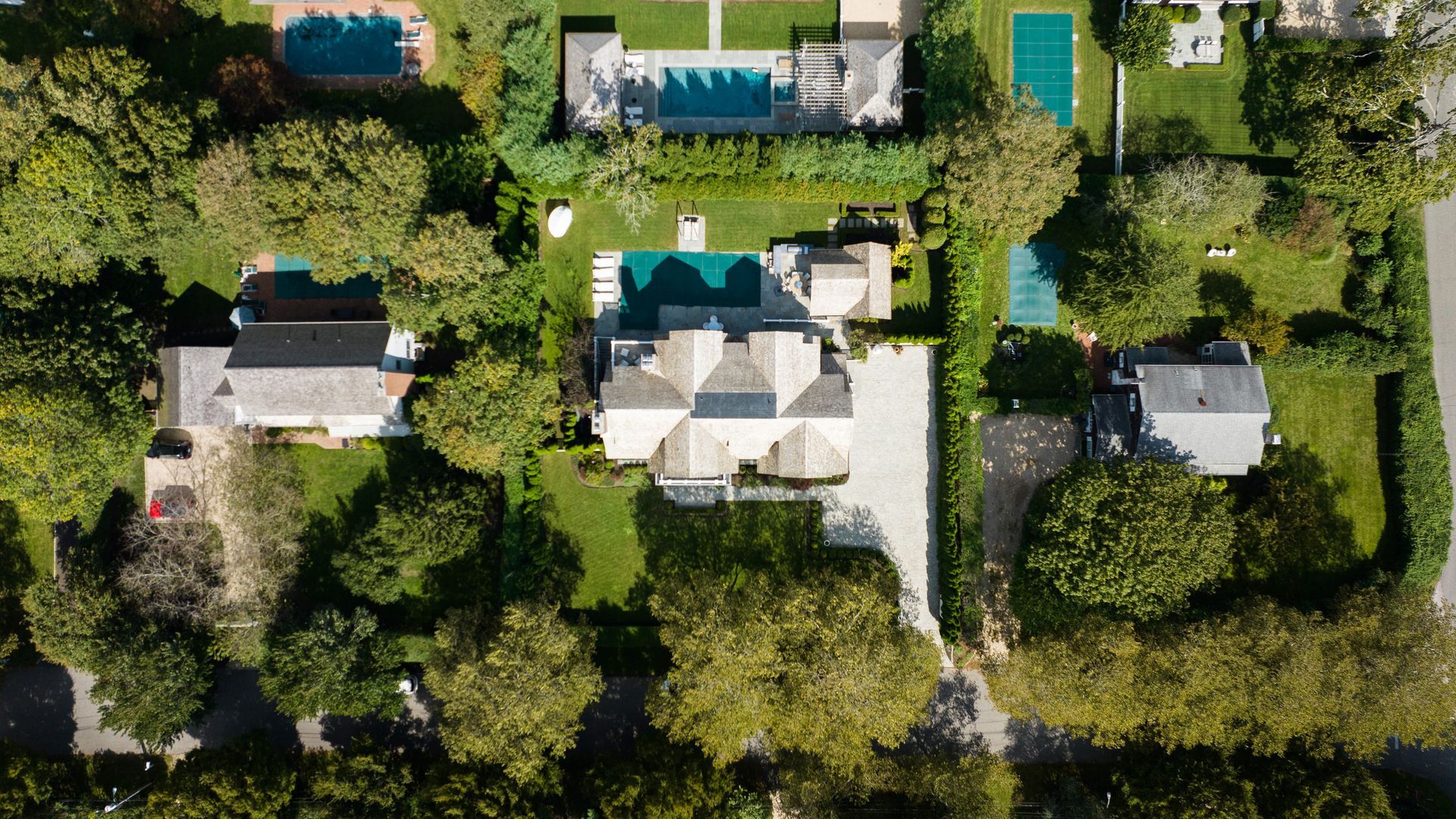 Aerial view of three suburban houses with swimming pools, lush green lawns, and mature trees on a sunny day.