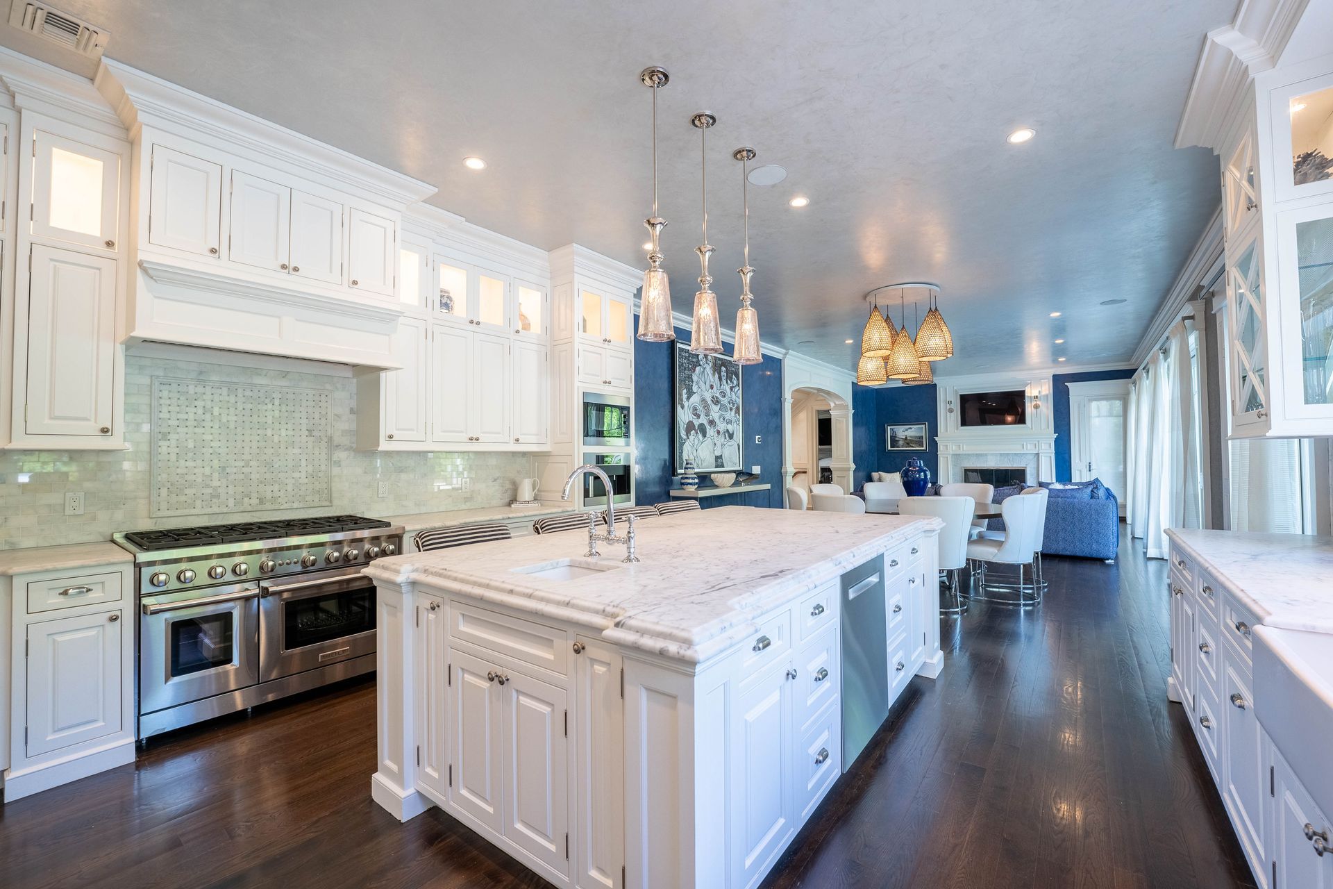 A white kitchen with marble countertops, a large center island, and stainless steel appliances over dark wood floors.