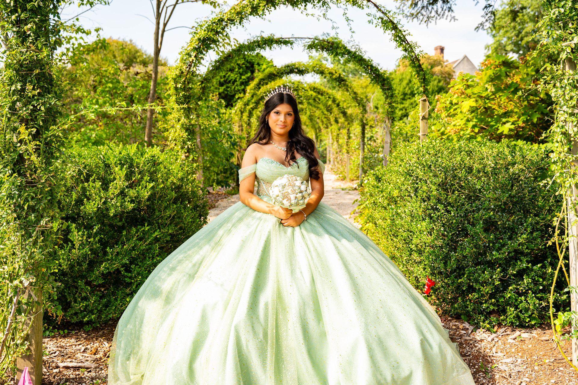 A person in a light green ball gown holding a bouquet stands under a long, arched ivy trellis in a sunlit garden.