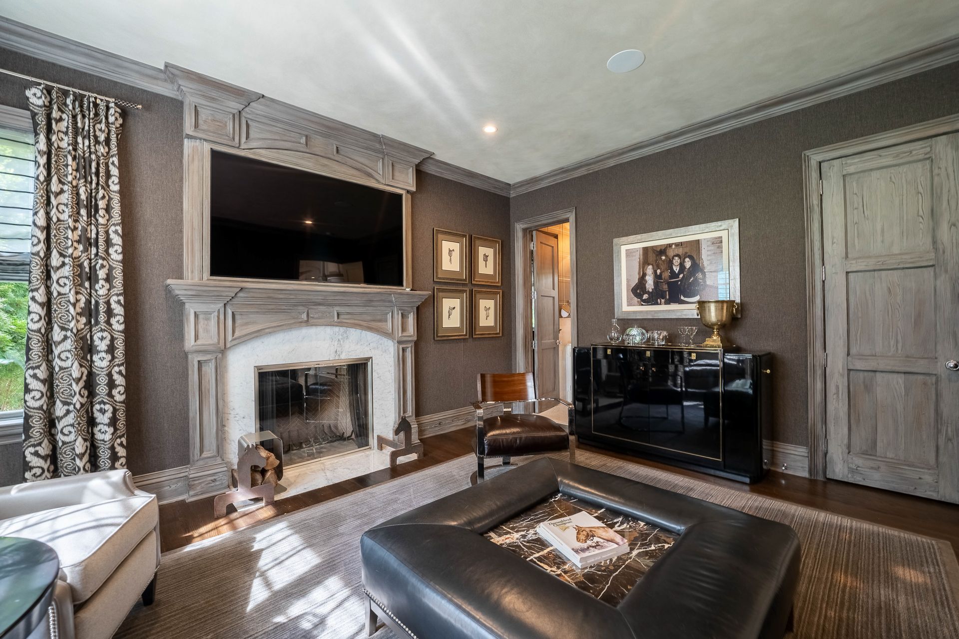 A living room featuring a fireplace with a mounted television, a leather ottoman, and dark patterned walls.