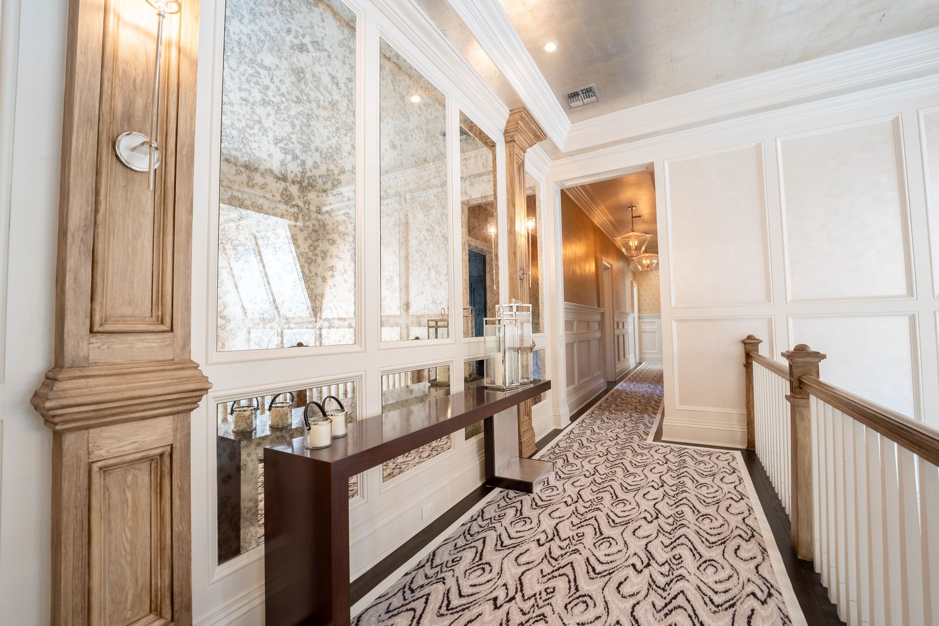A hallway with ornate wood trim, mirrored walls, a dark console table, and patterned carpet leading to another doorway.