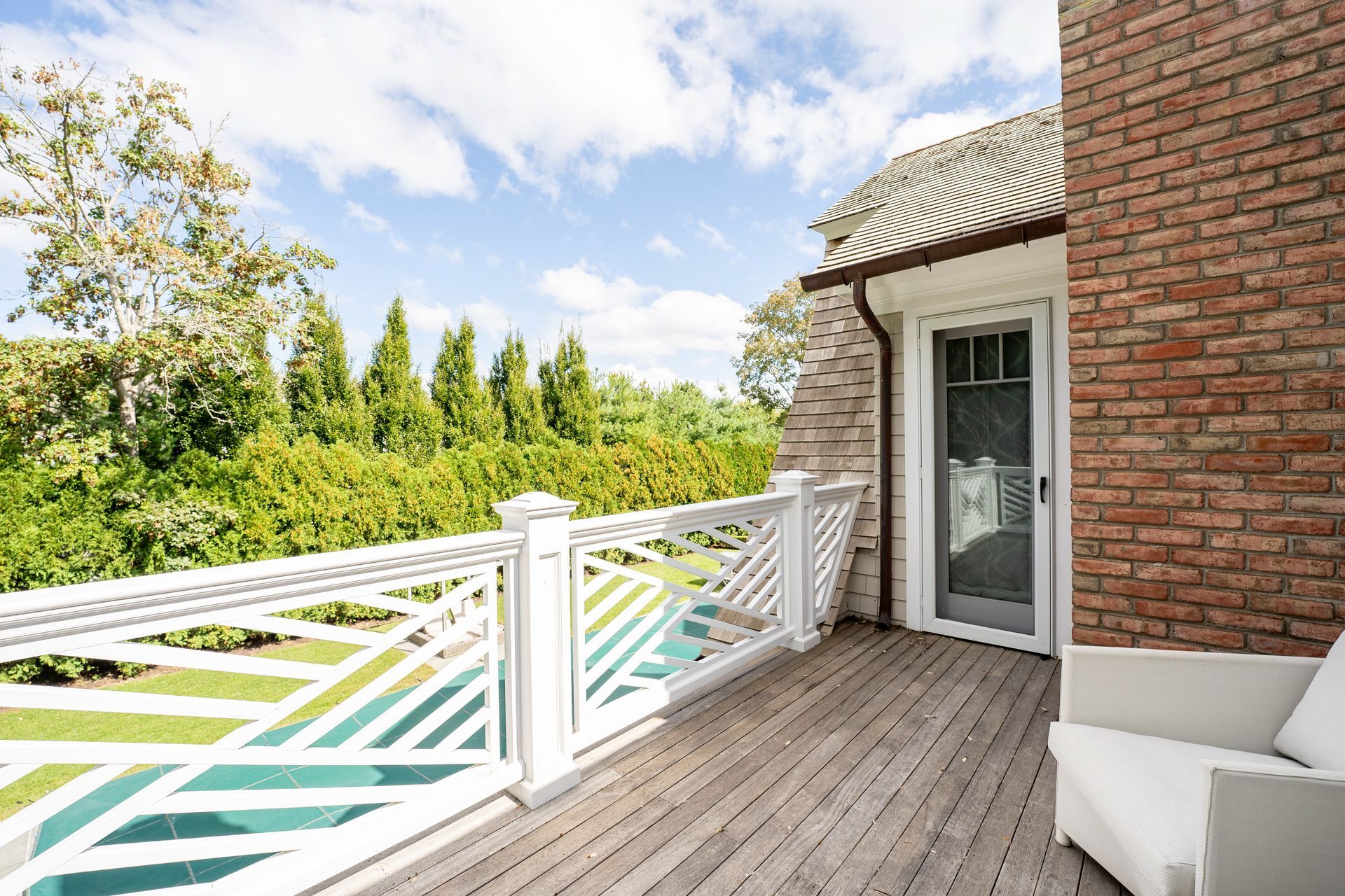 A wooden balcony with white railings overlooks a pool and garden next to a brick building.