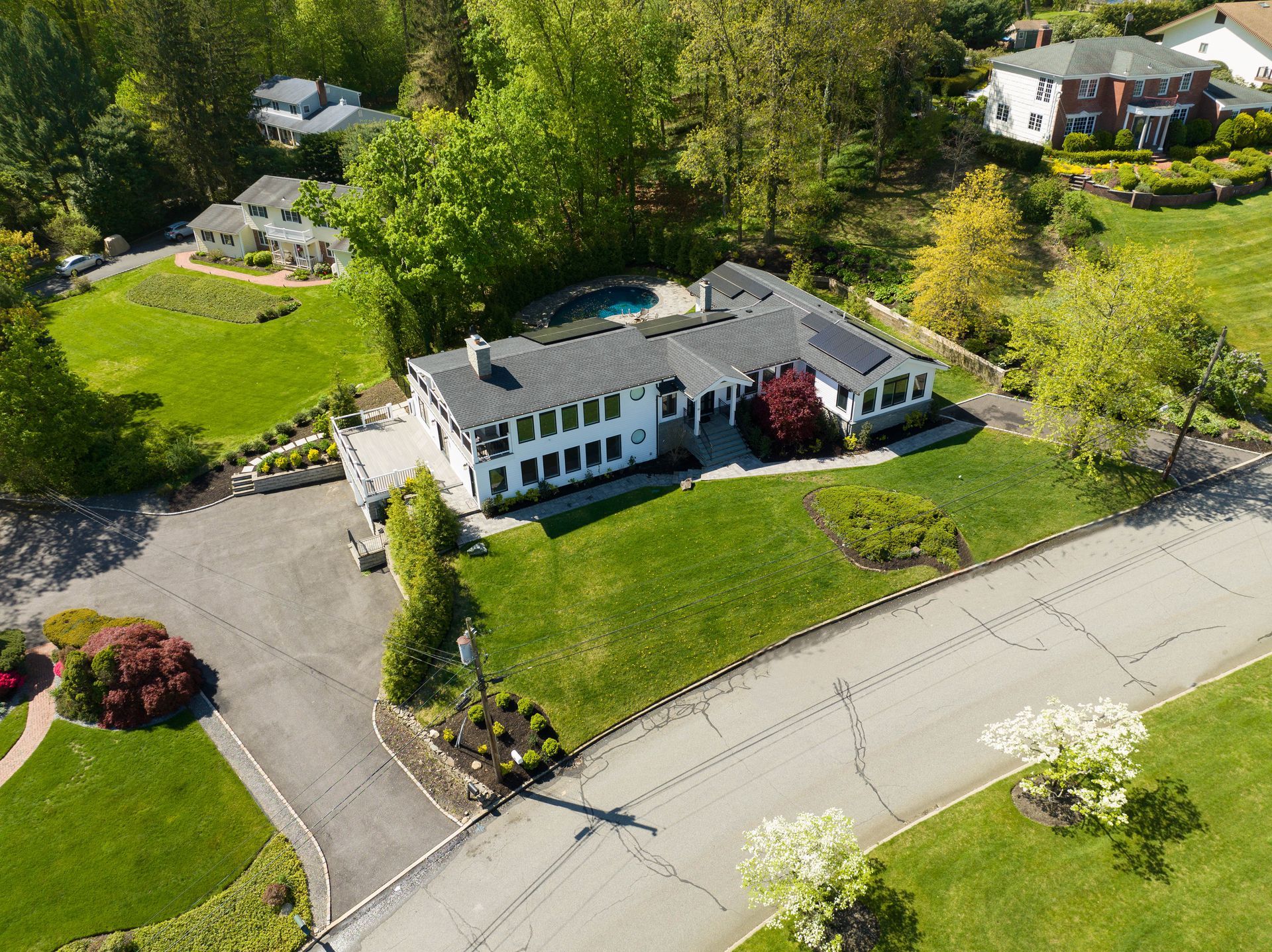 Aerial view of a white multi-story house with a grey roof, swimming pool, and circular driveway in a suburban area.