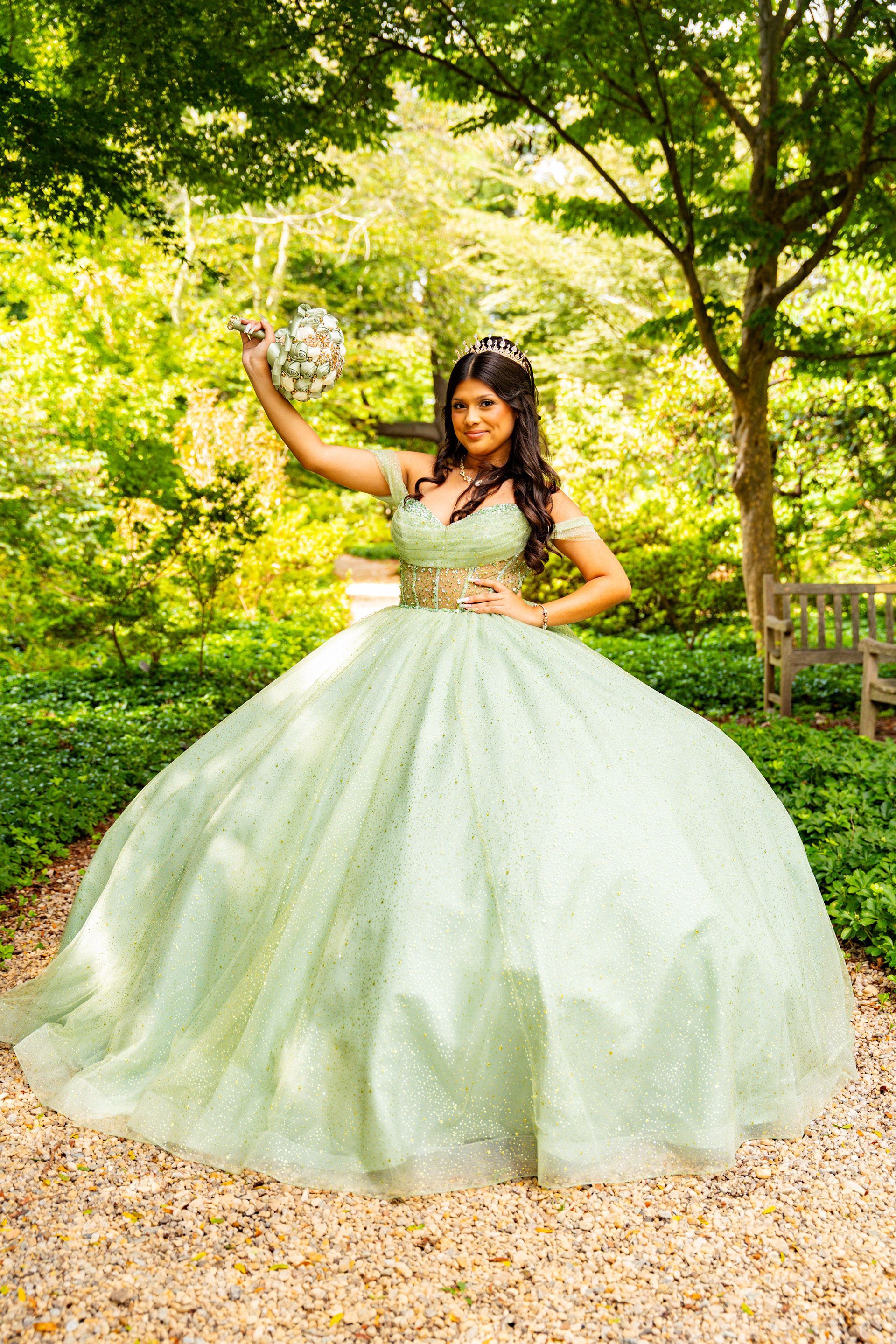 A person in a sparkling, light green ballgown and tiara stands in a garden, holding a bouquet of flowers aloft.