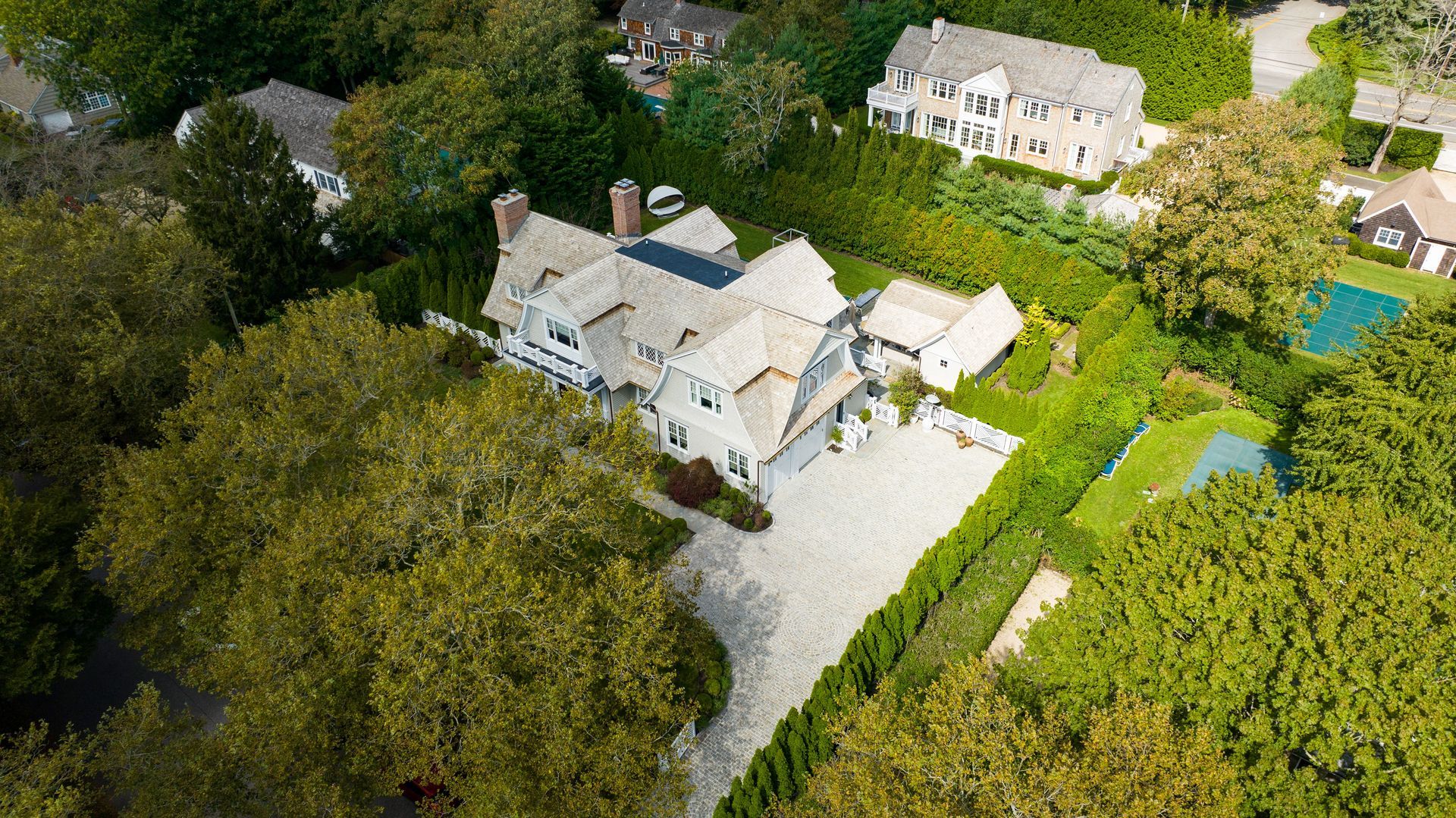 Aerial view of a white, multi-story shingle-style house with a gravel driveway, surrounded by lush green trees and lawns.