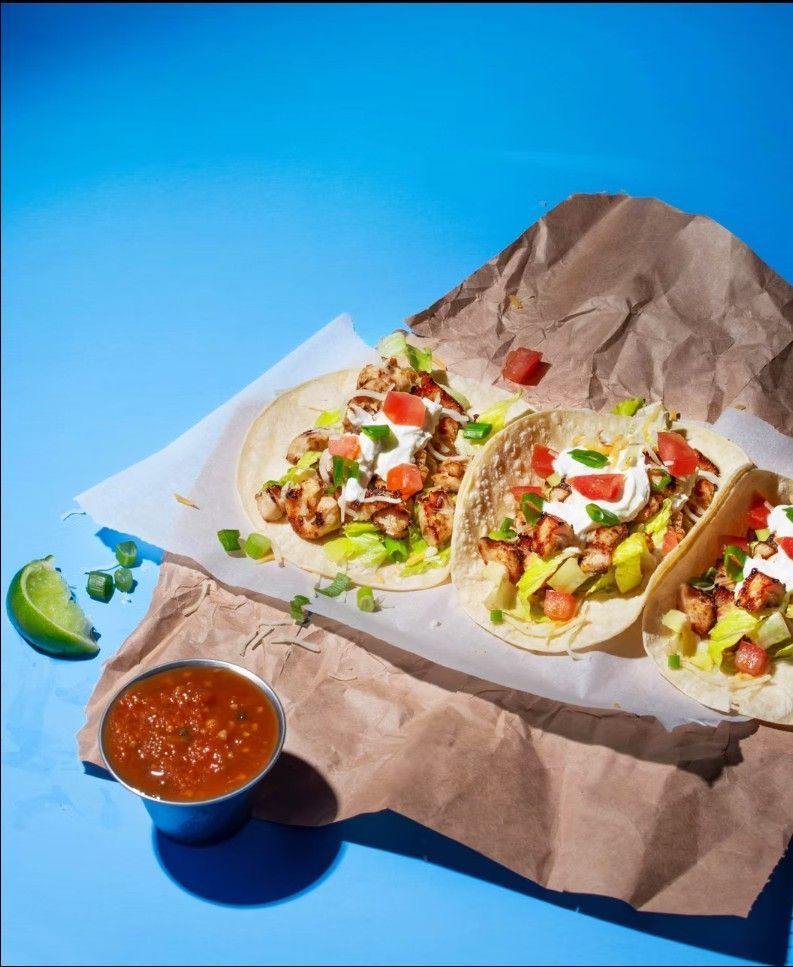 Three tacos topped with meat, lettuce, and sour cream on brown paper, with a side of salsa and a lime wedge on blue.