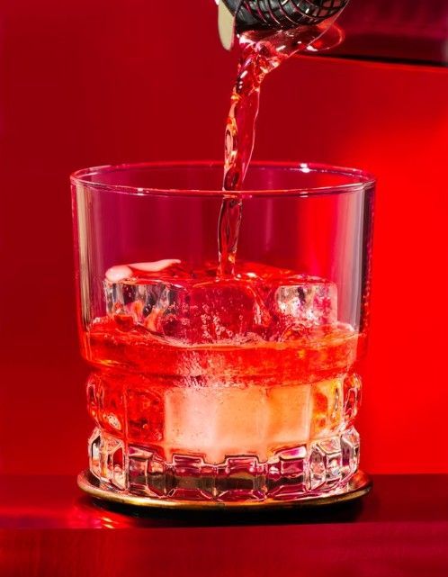 A bright red liquid is being poured into a textured glass filled with ice cubes against a solid red background.