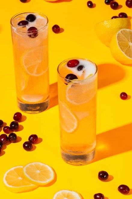 Two tall glasses of yellow citrus drink with cranberries, served on a bright yellow background with lemon slices.