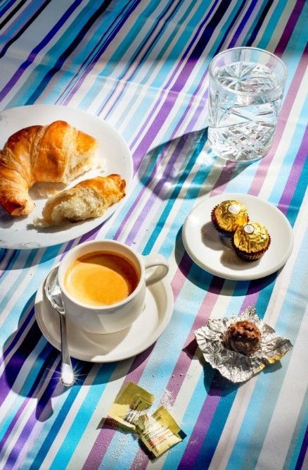 A breakfast spread with a croissant, coffee, a glass of water, and Ferrero Rocher chocolates on a blue-striped tablecloth.