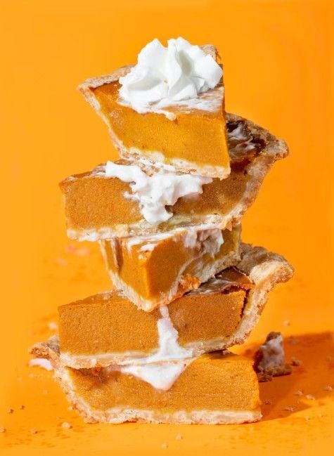 A vertical stack of four pumpkin pie slices topped with whipped cream against a solid orange background.