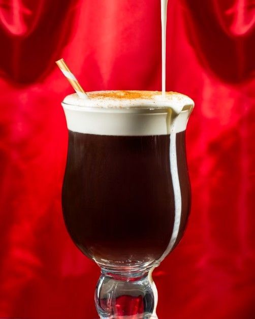 Cream being poured into an Irish coffee in a glass mug against a vibrant red backdrop.