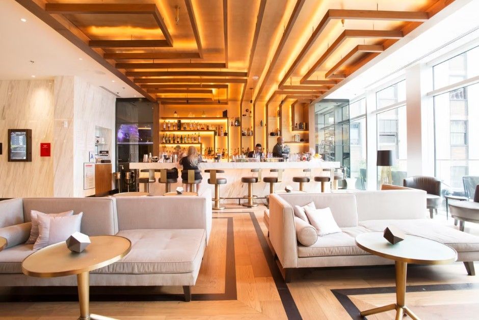 Modern hotel lobby with two light gray sofas, round coffee tables, and a bar area with wooden ceiling beams and warm lights.