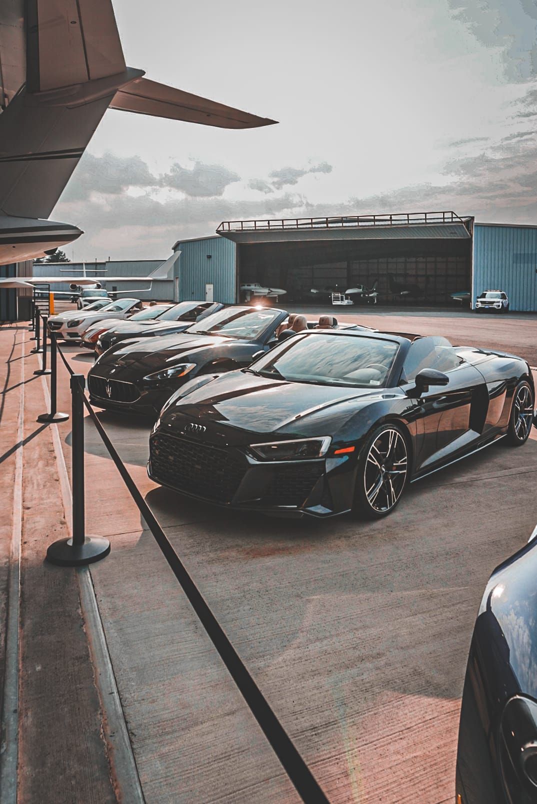 A lineup of luxury sports cars, including an Audi R8, parked on an airfield ramp near a hangar and an aircraft wing.