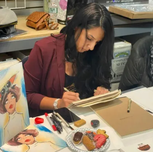 A person sits at a table drawing in a sketchbook, with artistic supplies and a small plate of pastries nearby.