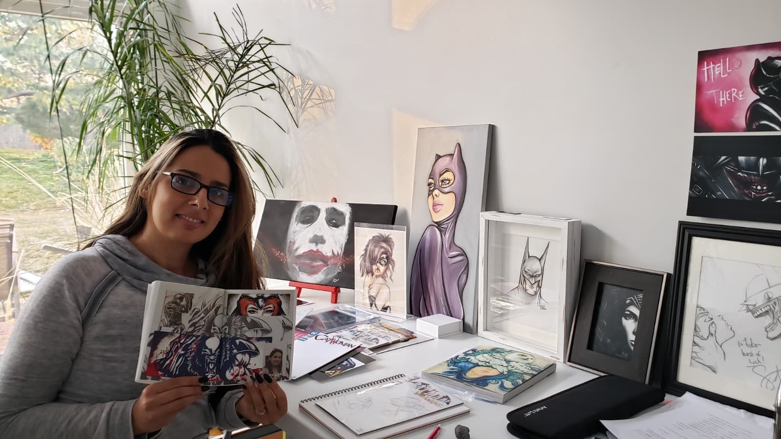 An artist holds a sketched drawing in a bright studio, surrounded by several Batman-themed character paintings and drawings.