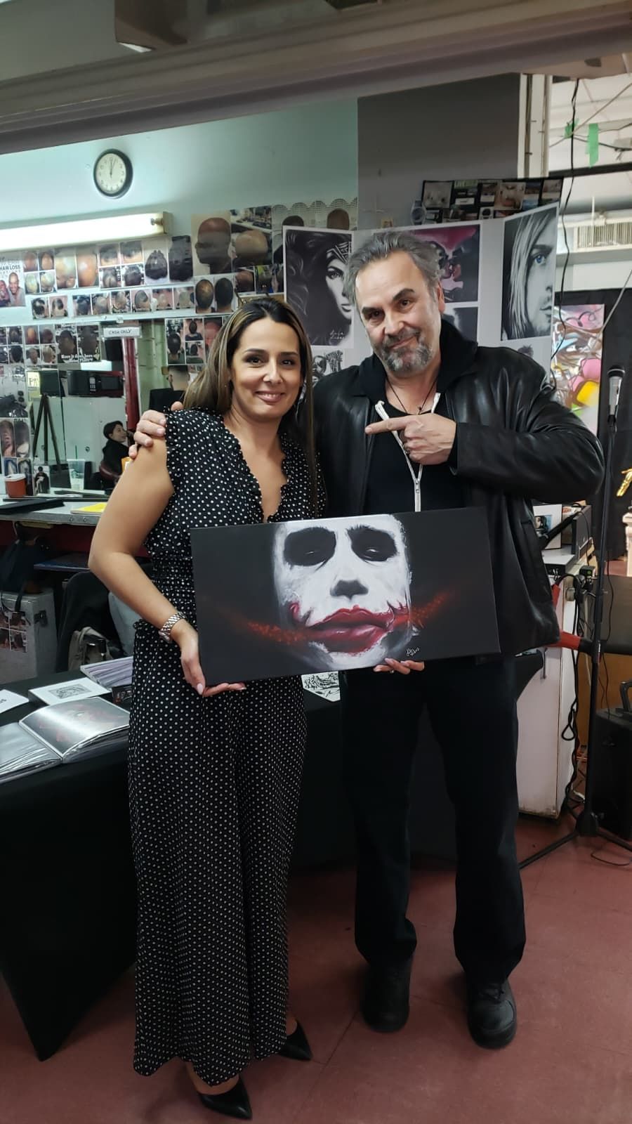 A smiling woman and a man in a leather jacket hold a canvas featuring a Joker-style mask in an art studio setting.
