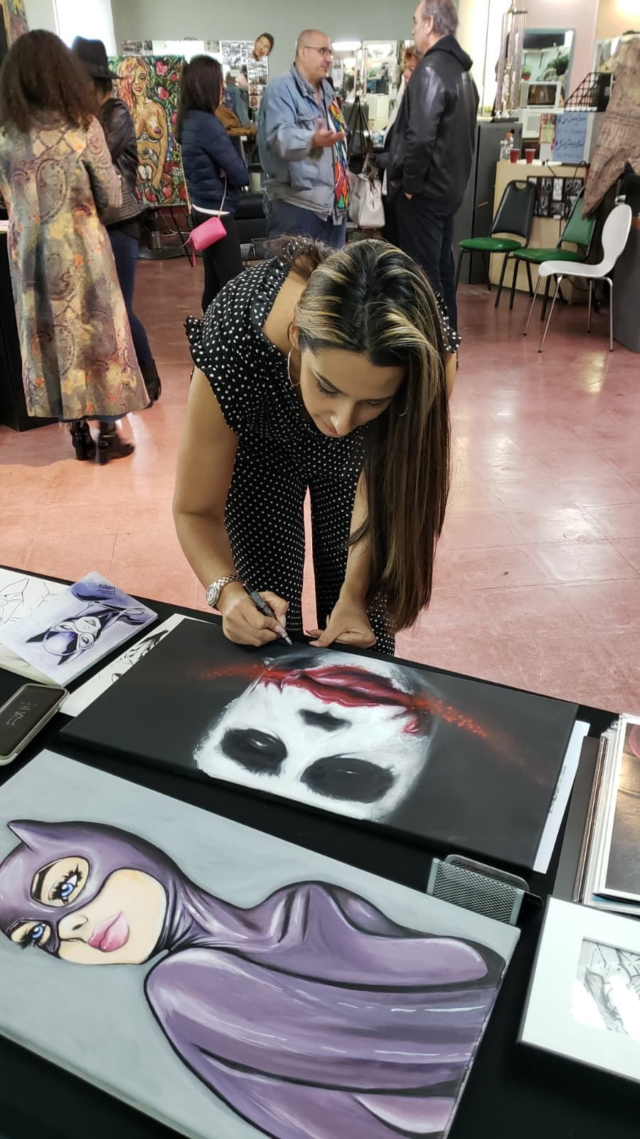 A person signs a painting of the Joker on a table next to another canvas depicting Catwoman in a crowded gallery space.
