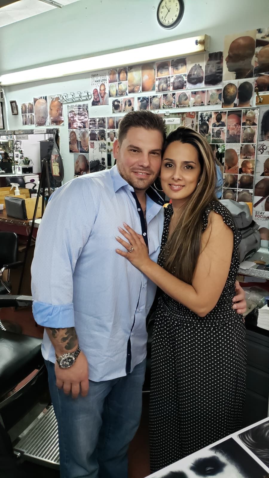 A man and woman smiling in a barbershop, standing side-by-side in front of a wall covered in hair style photos.