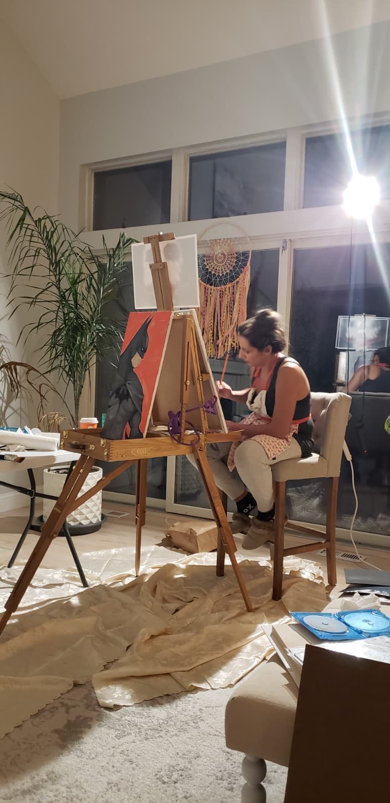A person painting on a canvas mounted on a wooden easel in a brightly lit room with a large potted plant nearby.