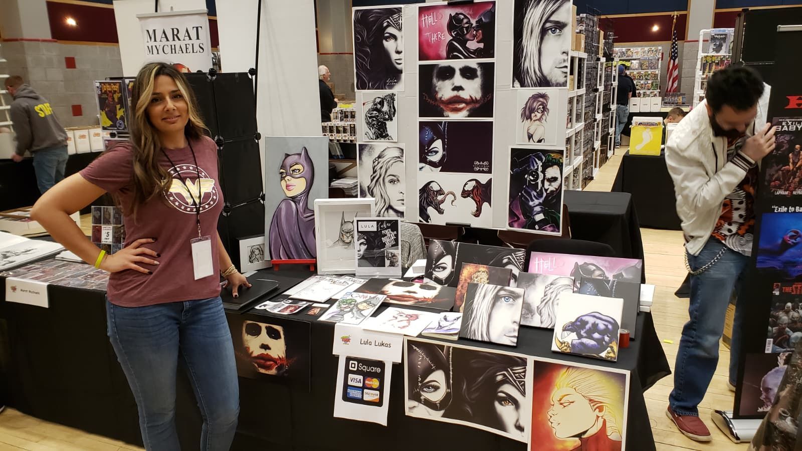 A smiling person stands at a booth filled with comic-style art prints at a convention, with a second person nearby.