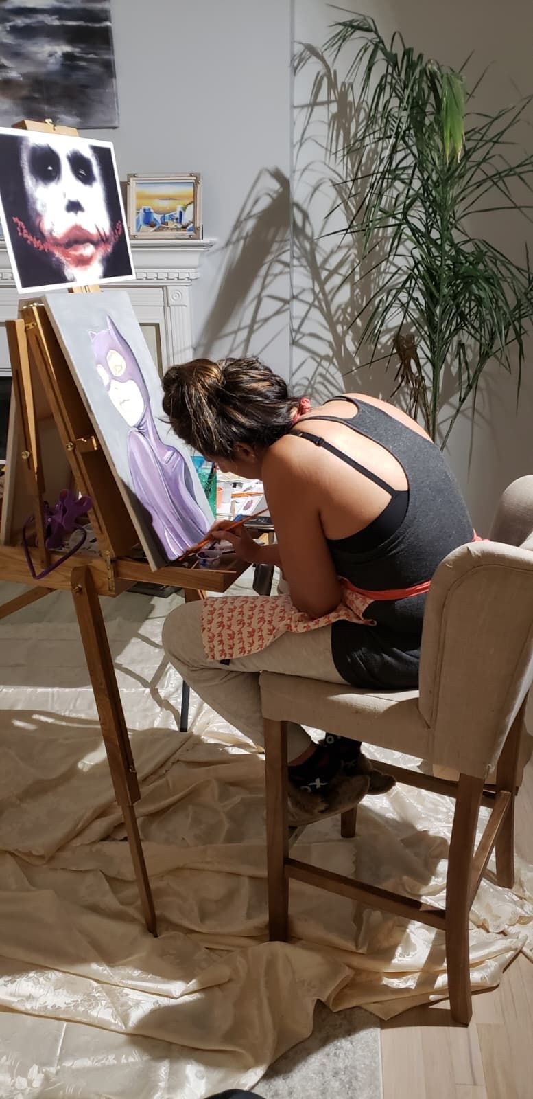 A person painting a portrait of the Joker on a canvas while seated at an easel in a room with a large potted plant.