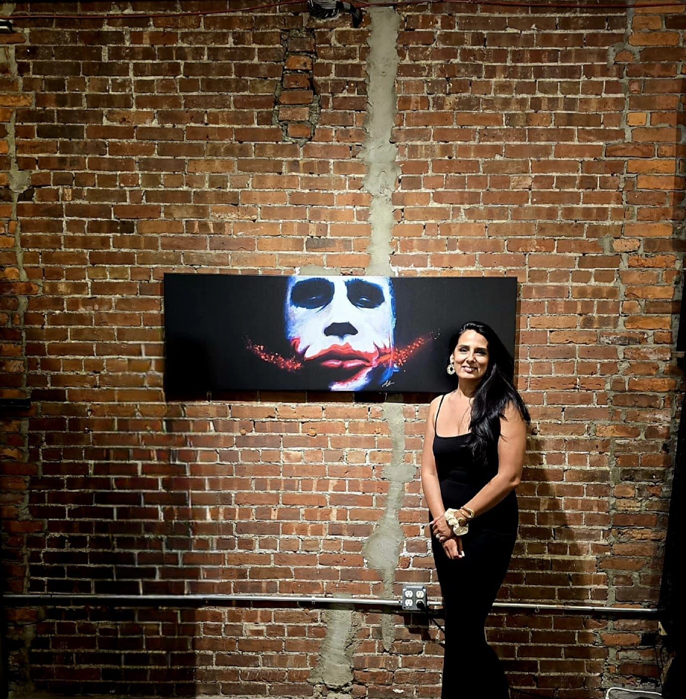 A person stands in a black outfit next to a rectangular painting of a clown-like face against a rustic brick wall.