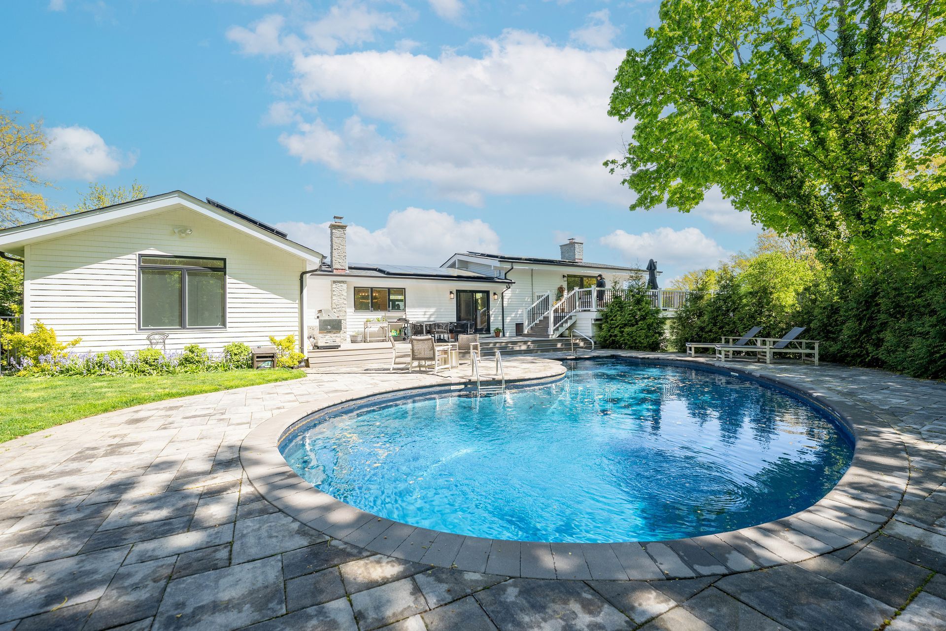 A sunny backyard featuring a rounded swimming pool, stone patio, white house, and lush green trees under a blue sky.