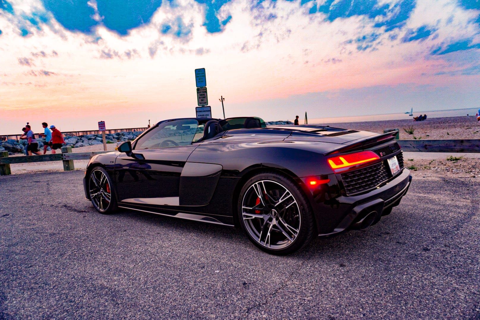 A black Audi R8 convertible parked on a gravel lot during a vibrant sunset near a beach with distant figures.