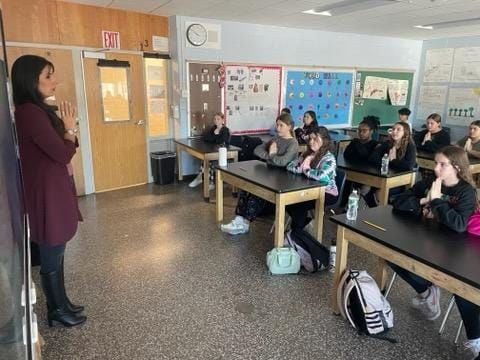 A teacher stands at the front of a classroom, hands pressed together, while students at desks mirror the gesture.