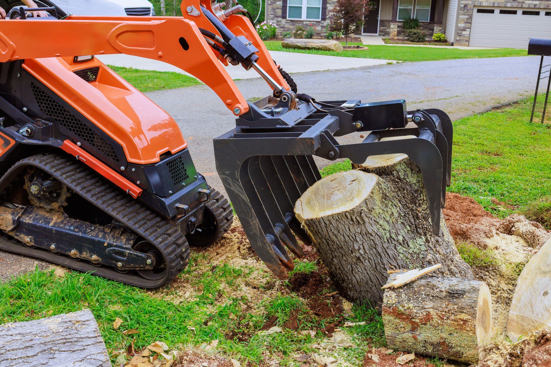 A loader lifts large tree logs with a grapple in a suburban yard.