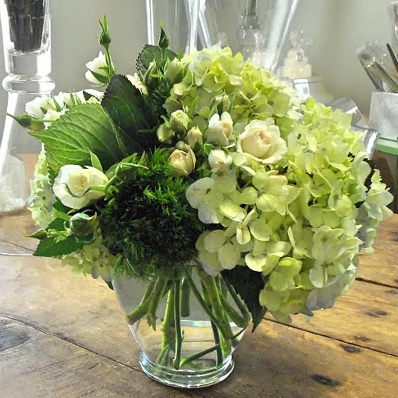A round glass vase holds an arrangement of white and green flowers, including hydrangeas and roses, on a wooden table.