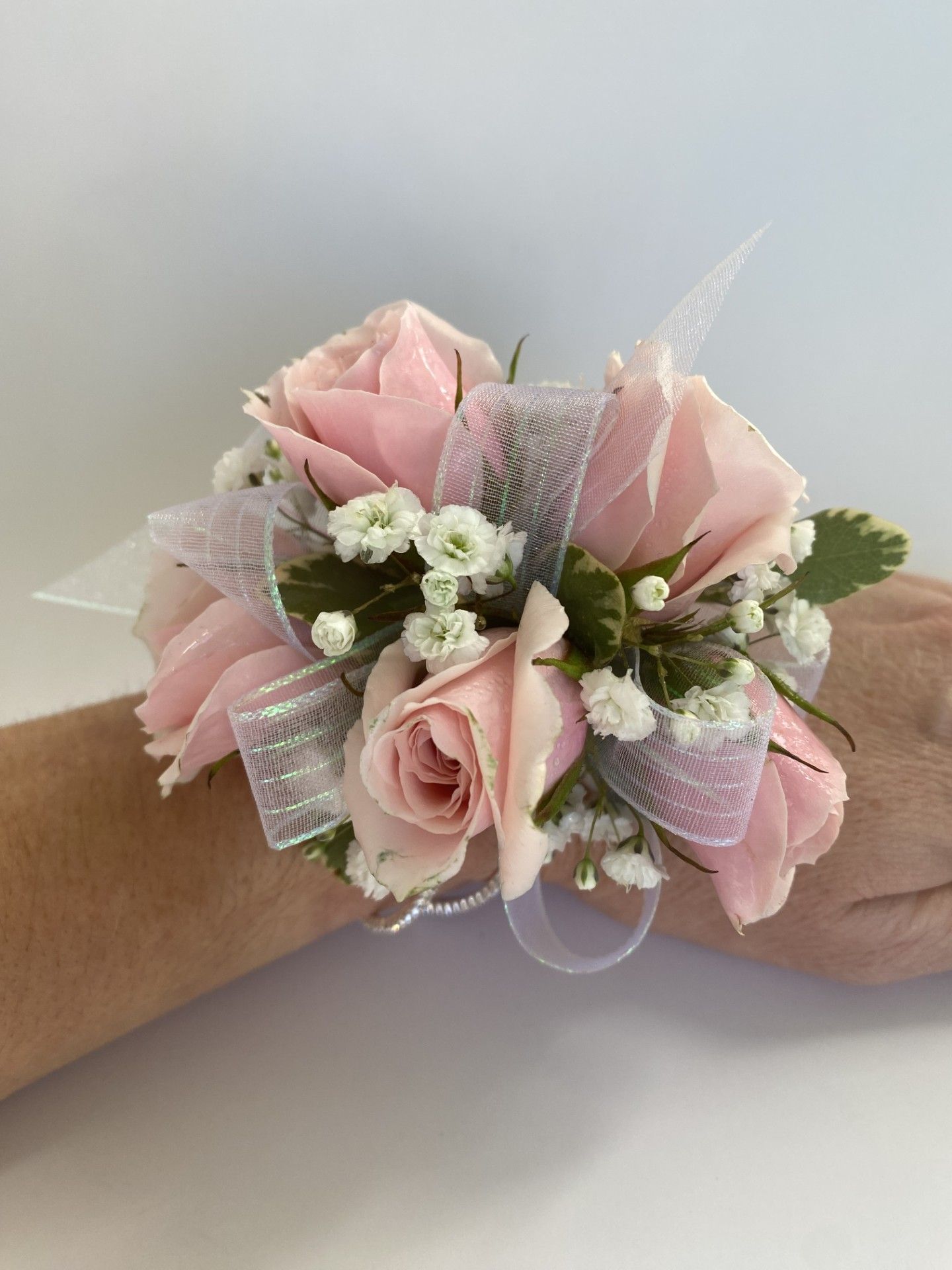 Wrist corsage with pink roses, white ribbons, and baby's breath on a light skin arm.