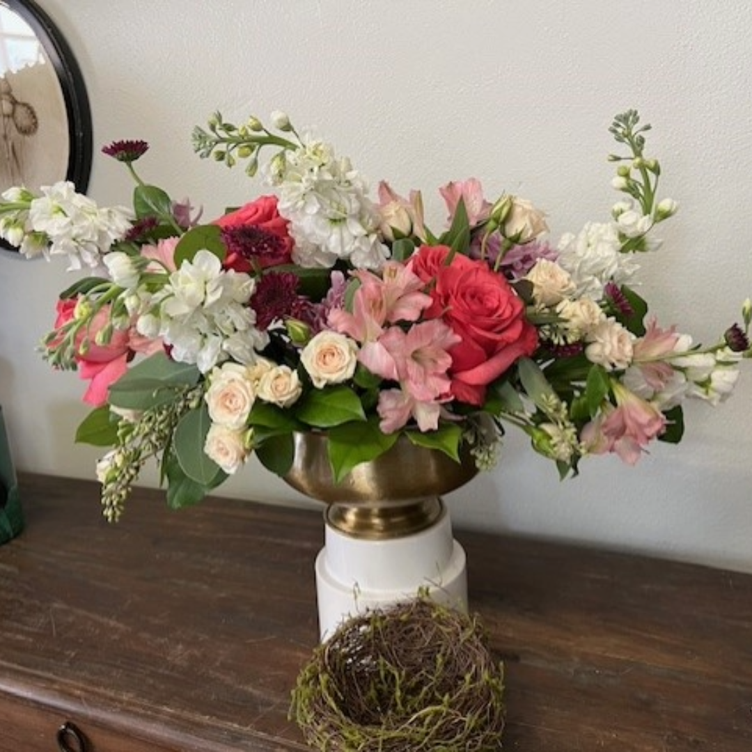 Floral arrangement in a gold vase on a white stand. Roses, snapdragons, and other flowers in pinks, white, and red.