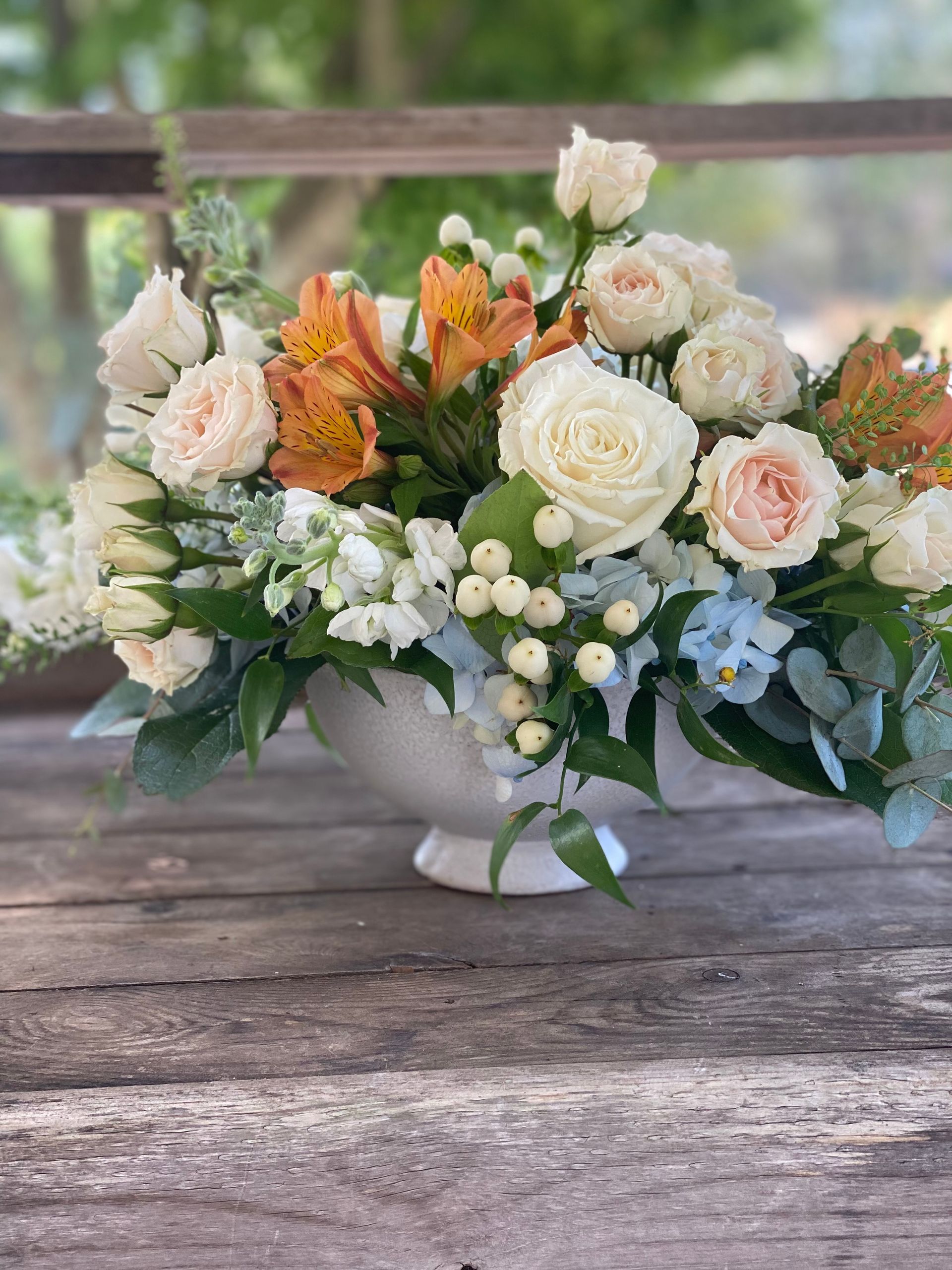 Floral arrangement in a white pedestal vase with orange, white, and blue flowers on a wooden table.