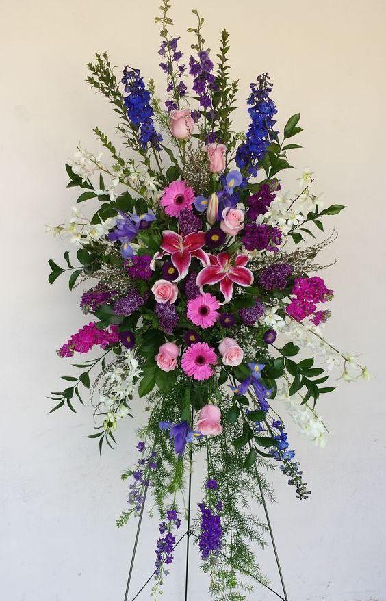 Funeral flower arrangement in purple, pink, blue, and white, on a metal stand.