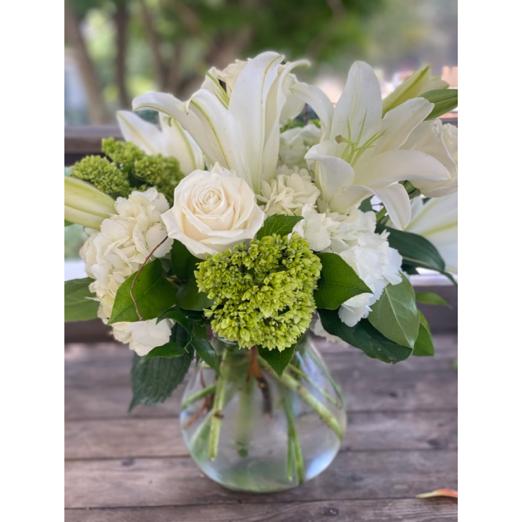 White flower bouquet in a clear glass vase on a wooden surface, with greenery and lilies.