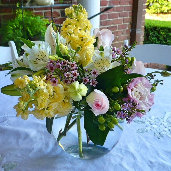 A vibrant bouquet of yellow, white, and pink flowers in a clear glass bowl on a white tablecloth.