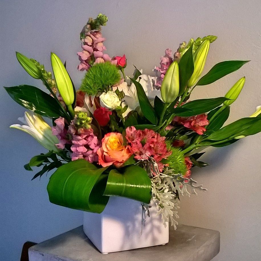 Colorful floral arrangement in a white square vase, with lilies, snapdragons, and other blooms.