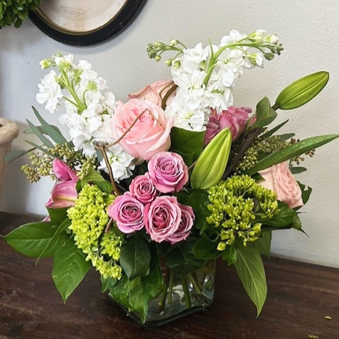 Floral arrangement in a square glass vase with pink and white roses, greenery, and lilies on a wooden table.