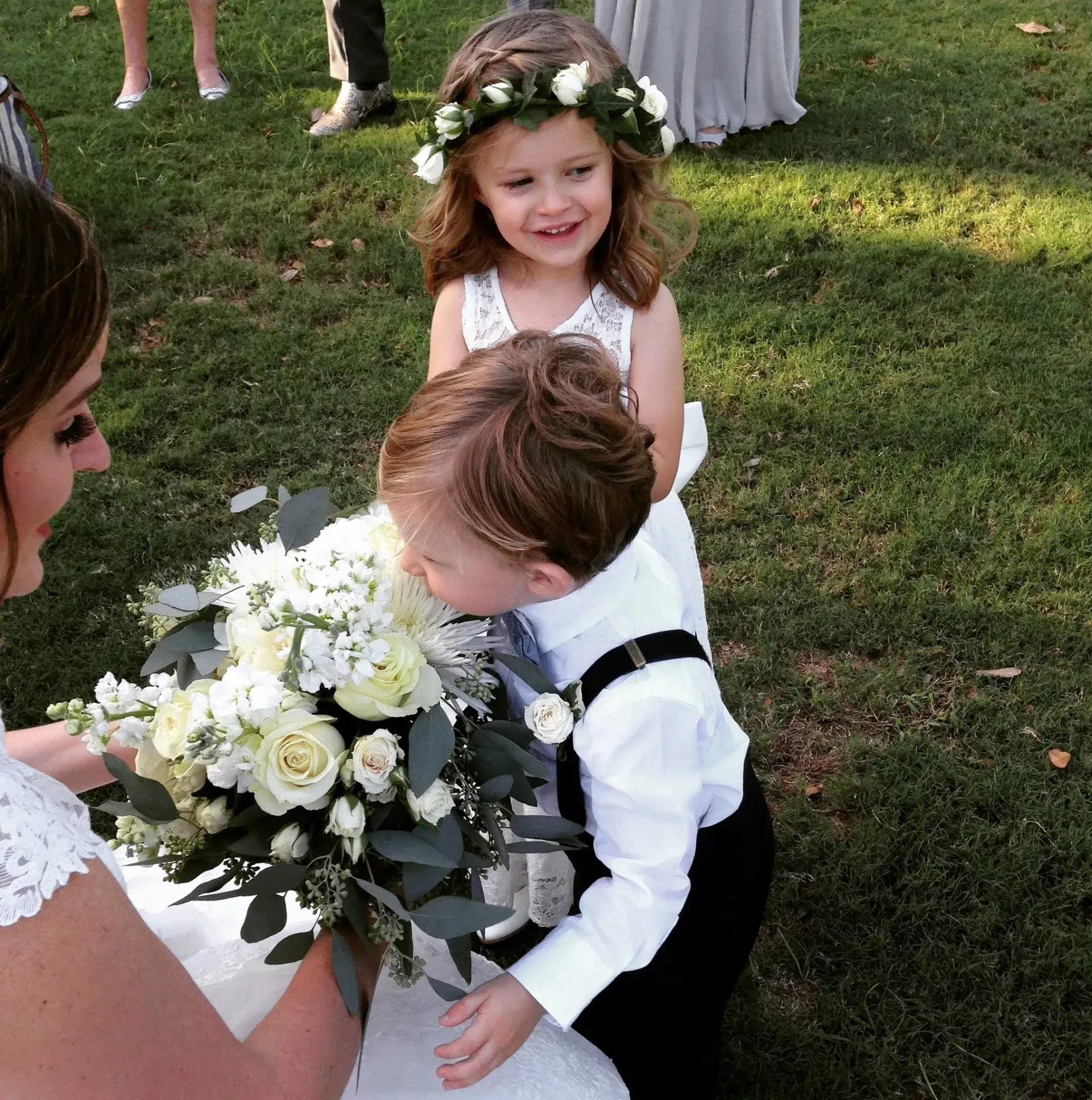 Bride with bouquet, flower girl in crown, ring bearer sniffing flowers at an outdoor wedding.