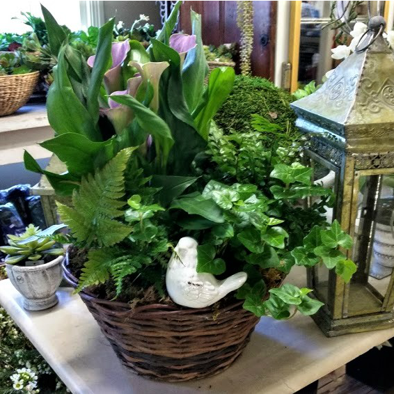 A basket overflowing with plants, calla lilies, ferns, and a white bird statue; set on a table.