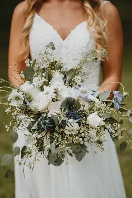 Bride in white dress holding a bouquet of white, blue, and green flowers outdoors.