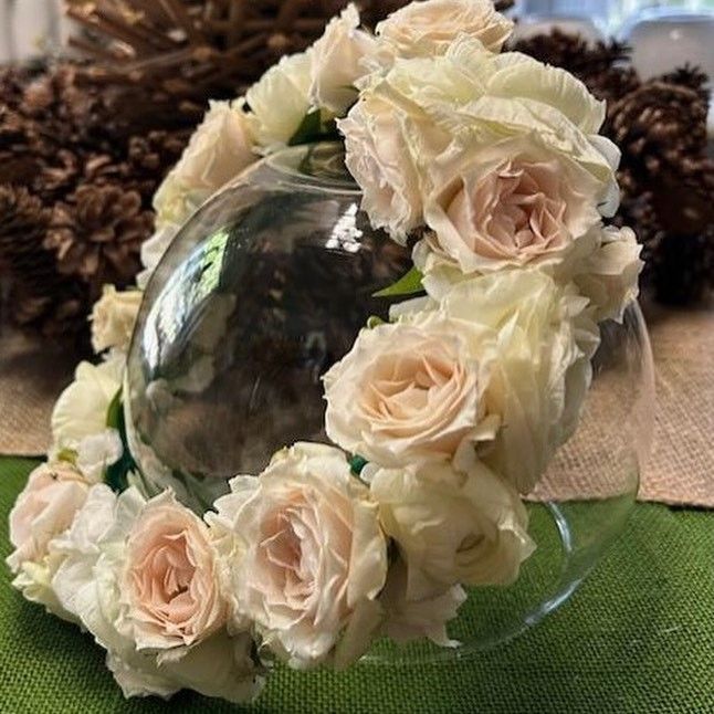 A clear glass bowl adorned with a ring of light peach roses sits on a green surface.