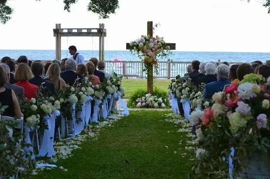 Wedding ceremony on a grassy area by the ocean; guests seated, awaiting the event; decorated with flowers and a cross.