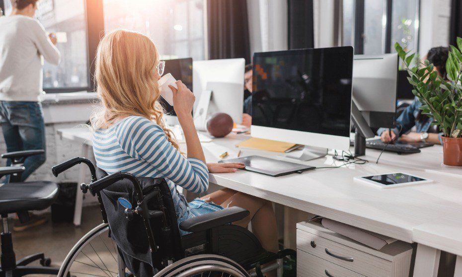 A woman in a wheelchair is at a desk in a share office looking at her screen while drinking