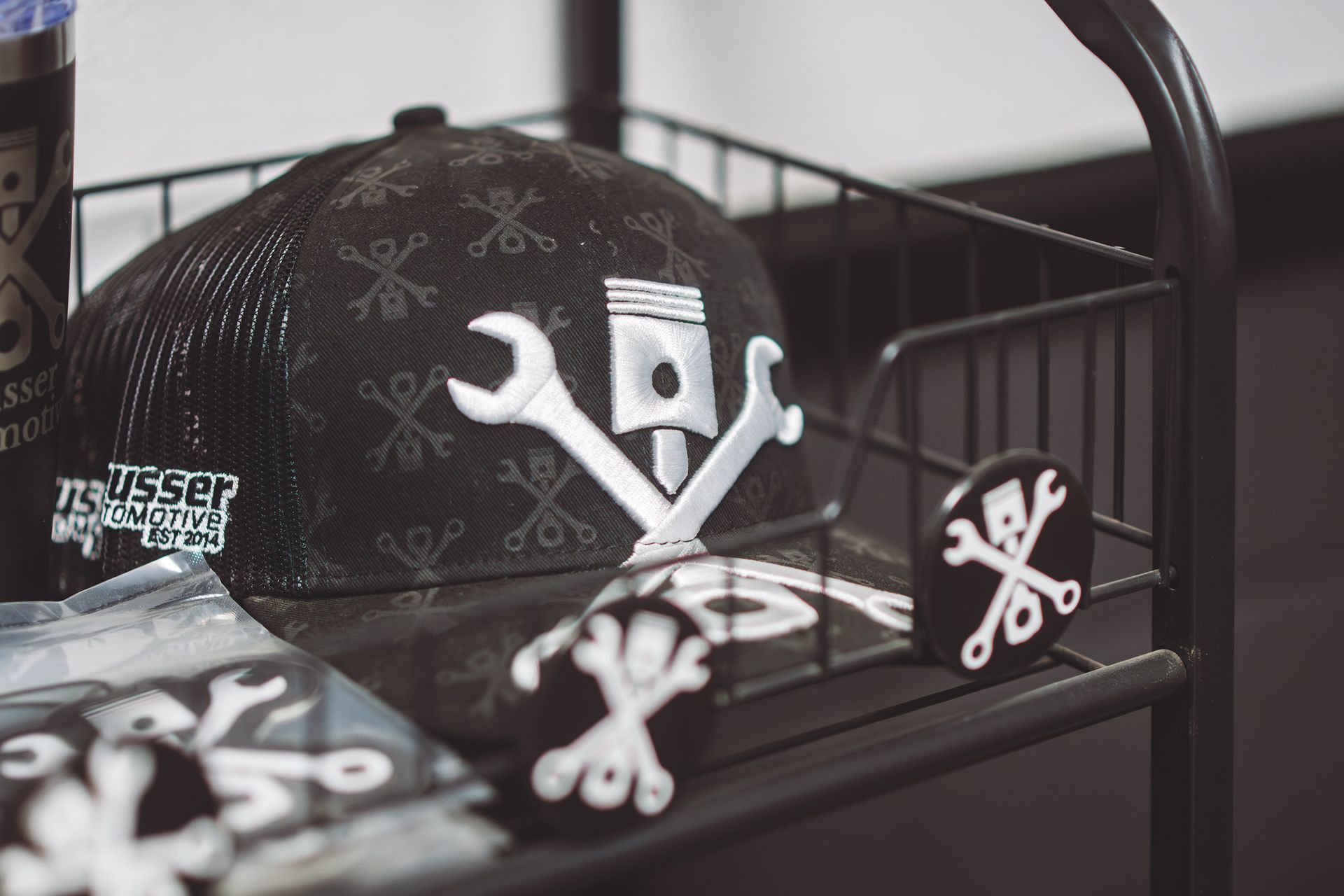 Black cap with white piston/wrench logo in a black metal basket with matching pins. | Musser Automotive