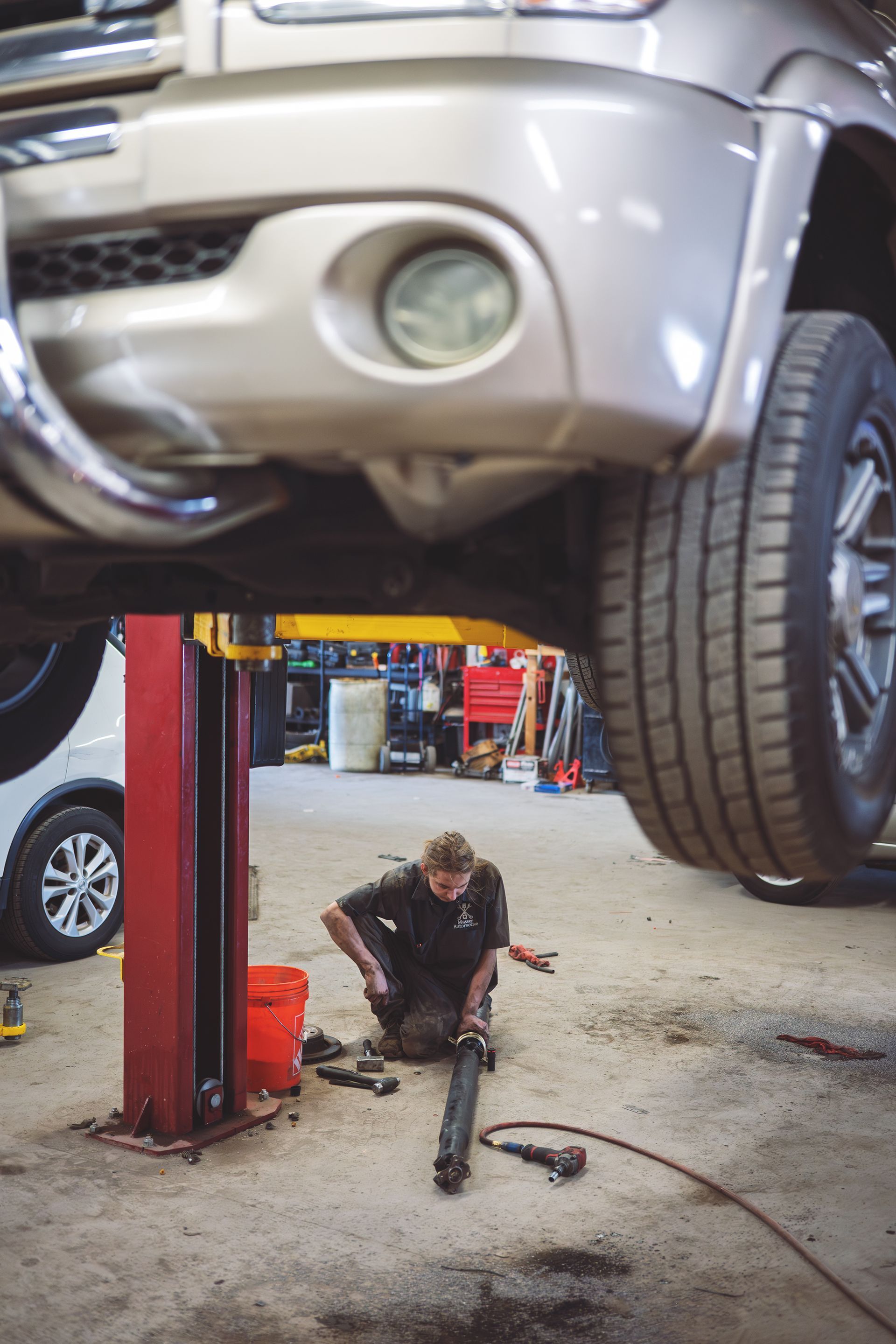 Mechanic working under a lifted vehicle in a garage, using tools. | Musser Automotive